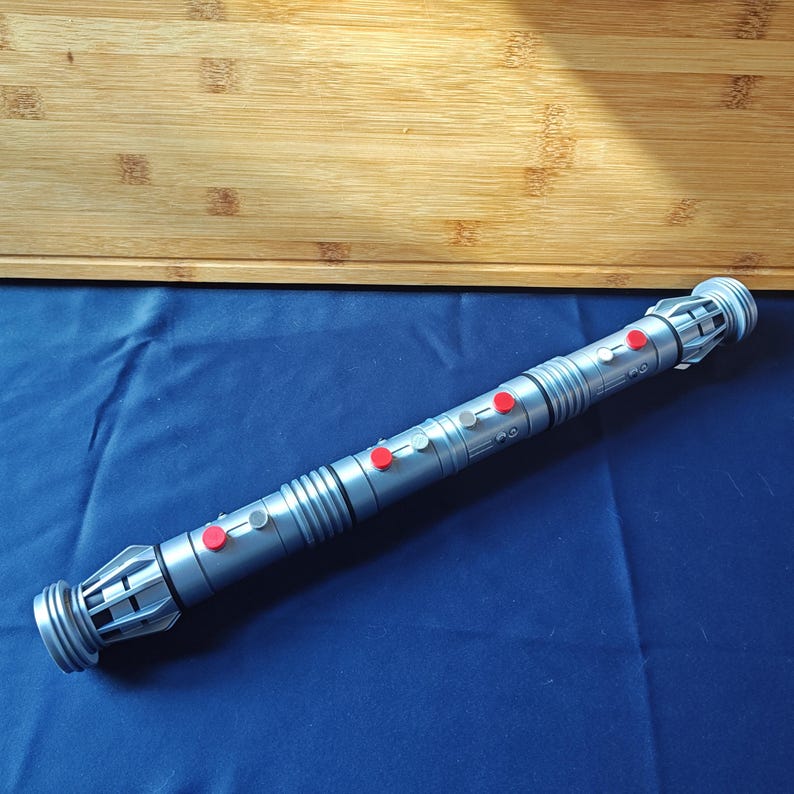 Puede incluir: Un accesorio de sable de luz plateado con botones rojos y grises. El accesorio tiene un cuerpo cil&iacute;ndrico con crestas y tapas de extremo detalladas. El sable de luz est&aacute; sobre una superficie azul, con una tabla de cortar de madera en el fondo.