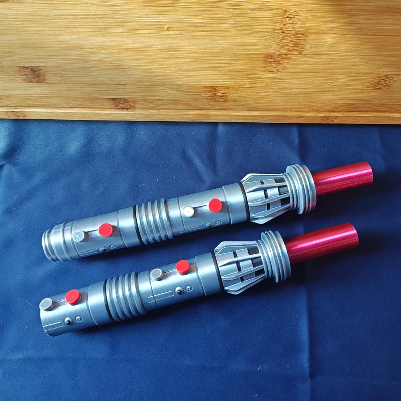 Puede incluir: Dos sables de luz de juguete plateados con detalles rojos y blancos. Los sables de luz tienen un dise&ntilde;o cil&iacute;ndrico con hojas rojas, sobre un fondo azul.