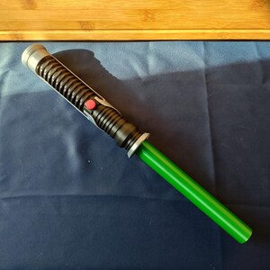 May include: A green and black lightsaber prop with a silver hilt and a red button. The lightsaber has a cylindrical black handle with ribbed detailing and a bright green blade. The prop is set against a dark blue background.