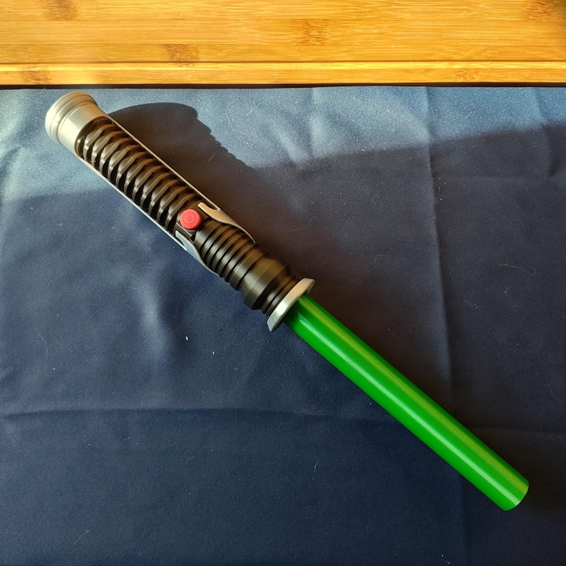 May include: A green and black lightsaber prop with a silver hilt and a red button. The lightsaber has a cylindrical black handle with ribbed detailing and a bright green blade. The prop is set against a dark blue background.