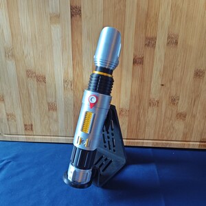 May include: A silver and black lightsaber prop with yellow and red accents, displayed on a black stand. The lightsaber has a cylindrical design with detailed ridges and buttons. The background is a wooden cutting board and a blue surface.