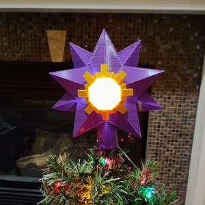 K&ouml;nnte beinhalten: Ein violetter, sternf&ouml;rmiger Christbaumspitze mit einem leuchtenden gelben Zentrum. Der Stern befindet sich oben auf einem gr&uuml;nen Weihnachtsbaum mit roten und gr&uuml;nen Lichtern.