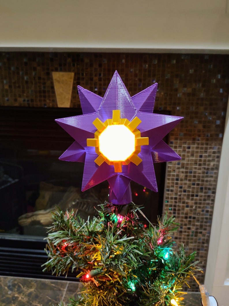 K&ouml;nnte beinhalten: Ein violetter, sternf&ouml;rmiger Christbaumspitze mit einem leuchtenden gelben Zentrum. Der Stern befindet sich oben auf einem gr&uuml;nen Weihnachtsbaum mit roten und gr&uuml;nen Lichtern.