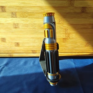 May include: A silver and gold lightsaber prop with a black handle. The lightsaber has a cylindrical design with gold accents and a black grip. The top features a gold ring and a pink gem.