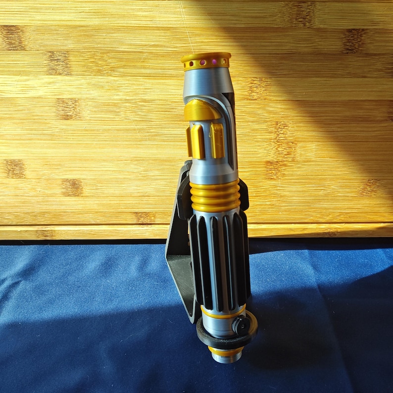 May include: A silver and gold lightsaber prop with a black handle. The lightsaber has a cylindrical design with gold accents and a black grip. The top features a gold ring and a pink gem.