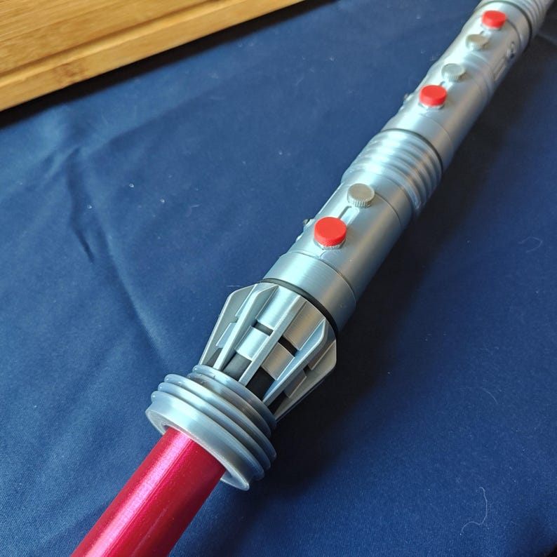 Puede incluir: Un sable de luz plateado y gris con botones rojos y grises. El mango tiene un dise&ntilde;o detallado con una hoja roja que se extiende desde la base. El sable de luz est&aacute; sobre un fondo azul.