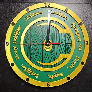 Puede incluir: Un reloj de pared redondo con esfera verde y detalles dorados. El reloj muestra las palabras "Second Breakfast", "Elevenses", "Luncheon", "Afternoon Tea", "Dinner" y "Supper". Las manecillas son negras y rojas.