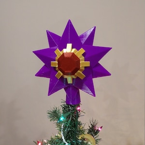 K&ouml;nnte beinhalten: Ein violetter, sternf&ouml;rmiger Christbaumspitze mit einem roten und goldenen Edelstein in der Mitte.