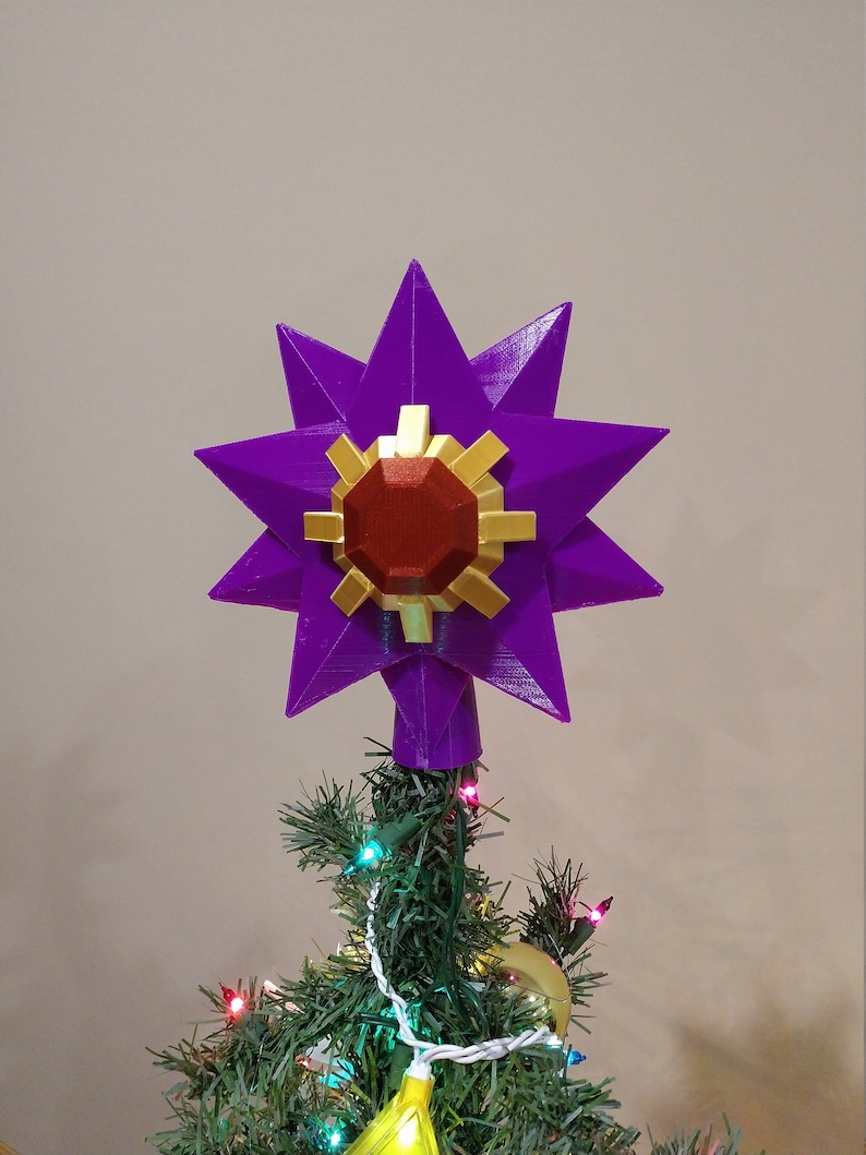 K&ouml;nnte beinhalten: Ein violetter, sternf&ouml;rmiger Christbaumspitze mit einem roten und goldenen Edelstein in der Mitte.
