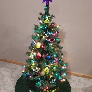 K&ouml;nnte beinhalten: Ein gr&uuml;ner Weihnachtsbaum, geschm&uuml;ckt mit Lichtern, Kugeln und einem Triforce-Baumrock. Der Baumspitzenstern ist lila mit einem roten Juwel in der Mitte.