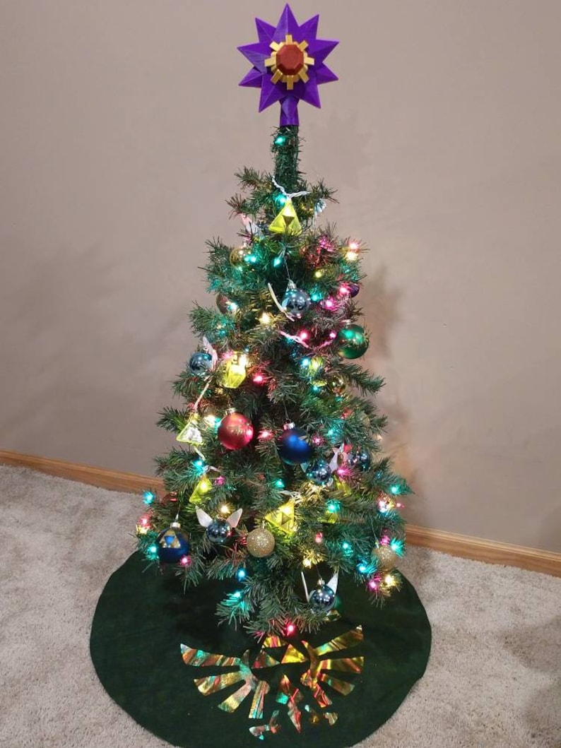 K&ouml;nnte beinhalten: Ein gr&uuml;ner Weihnachtsbaum, geschm&uuml;ckt mit Lichtern, Kugeln und einem Triforce-Baumrock. Der Baumspitzenstern ist lila mit einem roten Juwel in der Mitte.