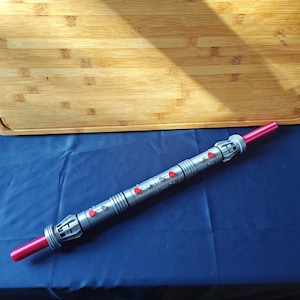 Puede incluir: Un accesorio de sable de luz plateado y rojo con un cuerpo cil&iacute;ndrico y detalles rojos. El sable de luz tiene una hoja roja en cada extremo y varios botones rojos a lo largo del cuerpo. El accesorio est&aacute; sobre una superficie azul.