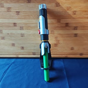 May include: A toy lightsaber with a black and silver handle, a red button, and a bright green blade. The lightsaber is resting in a black holder against a wooden surface and a blue surface.