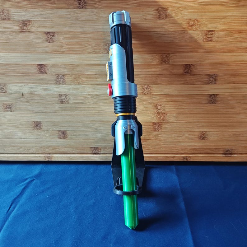 May include: A toy lightsaber with a black and silver handle, a red button, and a bright green blade. The lightsaber is resting in a black holder against a wooden surface and a blue surface.
