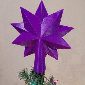 K&ouml;nnte beinhalten: Ein violetter, sternf&ouml;rmiger Christbaumspitzen. Der Stern ist aus einem 3D-gedruckten Material gefertigt und hat eine spitze Spitze.