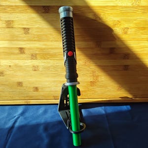 May include: A green and black lightsaber with a silver hilt and a red button. The lightsaber is mounted on a black holder. The background is a wooden surface and a blue surface.