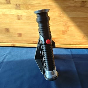 May include: A black lightsaber with a silver hilt and a red button, on a black stand. The lightsaber has a ribbed design and a grey top. The stand is on a blue surface.