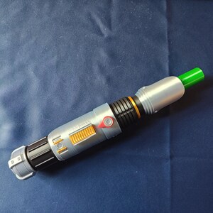 May include: A silver and black lightsaber prop with orange and red accents. The hilt features a detailed design with a green emitter. The prop is set against a dark blue background.