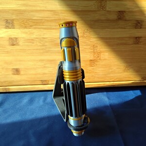 May include: A silver and gold lightsaber prop with a black stand. The lightsaber features gold accents and a ribbed handle. The top of the lightsaber has a gold ring with pink accents. The lightsaber is resting on a black stand.