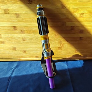 May include: A silver and yellow lightsaber with a purple blade, displayed on a black stand. The lightsaber has a ribbed handle and yellow accents. The blade is a solid purple color.