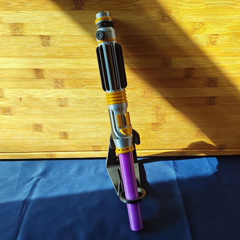 May include: A silver and yellow lightsaber with a purple blade, displayed on a black stand. The lightsaber has a ribbed handle and yellow accents. The blade is a solid purple color.