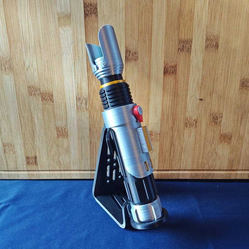 May include: A silver and black lightsaber with a black stand. The lightsaber has a silver hilt with black and yellow accents, a red button, and a silver blade emitter. The stand is black and triangular, providing support for the lightsaber.