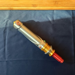 May include: A silver and gold lightsaber prop with red and green accents. The hilt has a detailed design with a red emitter. The lightsaber is lying on a dark blue surface.