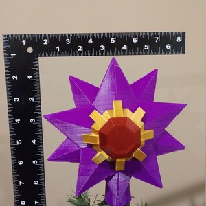 K&ouml;nnte beinhalten: Ein violetter, sternf&ouml;rmiger Christbaumspitze mit einem roten und goldenen Edelstein in der Mitte. Die Spitze ist etwa 15 cm hoch.