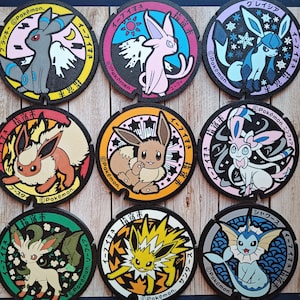 Puede incluir: Nueve posavasos coloridos con temática Pokémon. Cada posavasos presenta un personaje Pokémon diferente en un diseño circular con texto japonés y el logotipo de Pokémon. Los posavasos son negros con acentos coloridos.