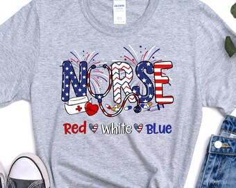 Puede incluir: Una camiseta gris con un gráfico rojo, blanco y azul que dice "NURSE" con un estetoscopio, un corazón y otros símbolos médicos. El texto "Red White Blue" está debajo del gráfico.