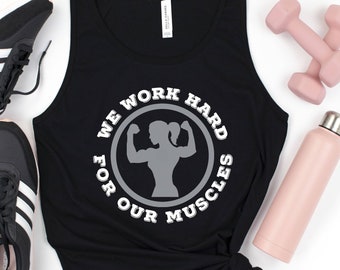 May include: Black tank top with a white circle graphic that says "We work hard for our muscles" with a silhouette of a woman lifting weights inside the circle. The tank top is laying on a white surface with a water bottle, two dumbbells, and two black sneakers.