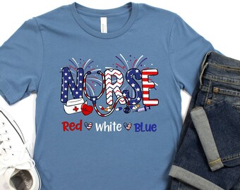 Puede incluir: Una camiseta azul con un gráfico de la palabra "NURSE" en rojo, blanco y azul con un estetoscopio y otros símbolos médicos. El texto "Red White Blue" está debajo del gráfico.
