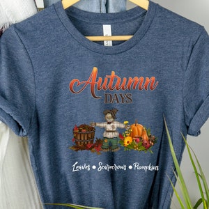 May include: A navy blue t-shirt with the text "Autumn Days" in orange and yellow lettering. A cartoon scarecrow is holding a basket of leaves and standing next to a pumpkin. The text "Leaves, Scarecrows, Pumpkins" is below the image.