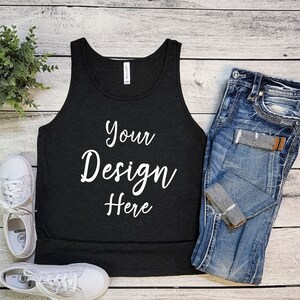 Pode incluir: Um top preto de alças com o texto "Your Design Here" impresso em branco sobre um fundo de madeira. O top está colocado plano com um par de calças de ganga azuis e ténis brancos ao lado.