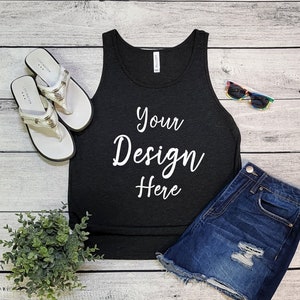 Pode incluir: Camisola de alças preta com o texto "Your Design Here" sobre um fundo de madeira branca. A camisola de alças é estilizada com calções de ganga, óculos de sol e sandálias.