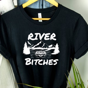 Pu&ograve; includere: Maglietta nera con testo bianco che recita "River Bitches" e una grafica di una catena montuosa con una barca su un lago.