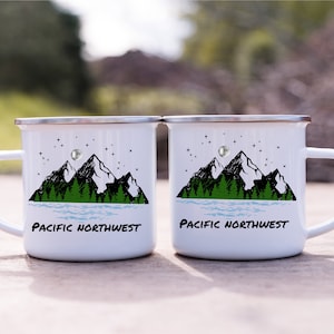 May include: Two white enamel mugs with black and green mountain and lake designs. The mugs have silver rims and handles. The text "Pacific Northwest" is printed on each mug.