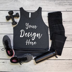 May include: A gray tank top with the text "Your Design Here" on a white background. The tank top is laying on a white wooden surface with a pair of black leggings, a black water bottle, and a black dumbbell.