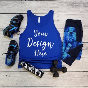 May include: A royal blue tank top with the text "Your Design Here" in white. The tank top is laid flat on a white wood surface with a pair of blue and black running shoes, a black and white water bottle, and a pair of black dumbbells.