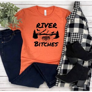 May include: Orange t-shirt with black text that reads "River Bitches" and a graphic of a mountain scene with a boat and a pumpkin.