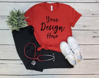 May include: Red t-shirt with the text "Your Design Here" on a white wood background. The t-shirt is paired with black pants, a stethoscope, and white sneakers.