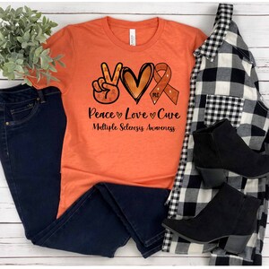 Puede incluir: Camiseta naranja con un signo de paz, un coraz&oacute;n y una cinta con el texto "Peace Love Cure" y "Multiple Sclerosis Awareness".