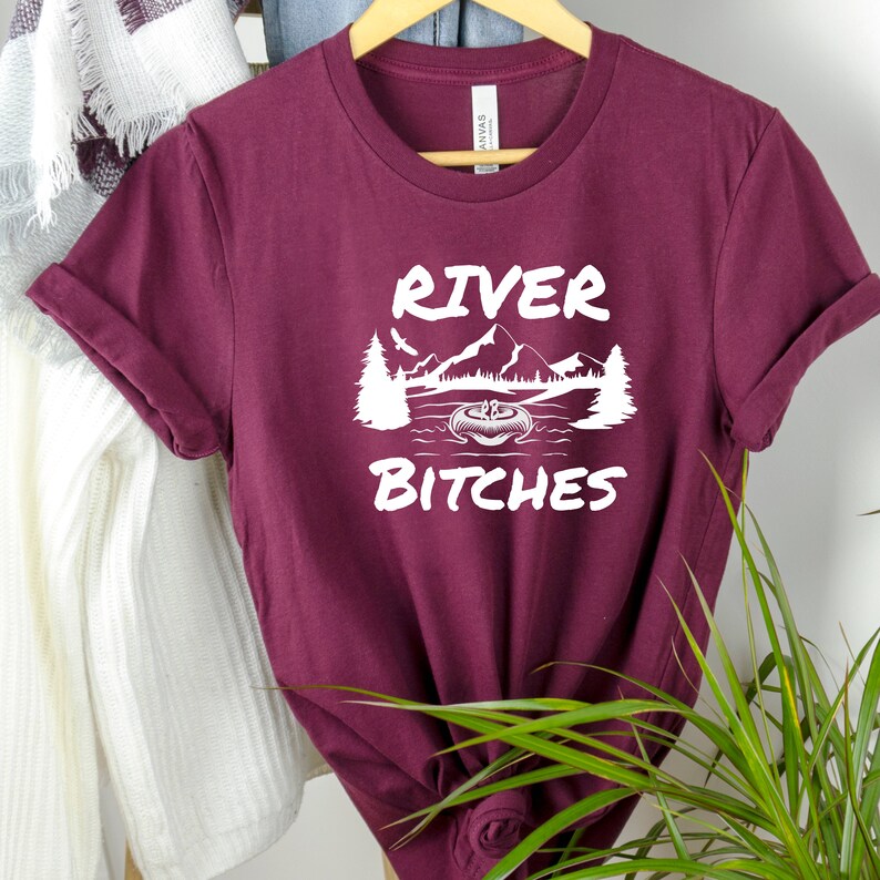 Pu&ograve; includere: Maglietta color borgogna con testo bianco che recita "River Bitches" e una grafica di una canoa in un lago con montagne sullo sfondo.