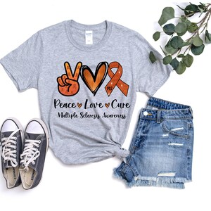Puede incluir: Camiseta gris con un signo de paz, un coraz&oacute;n y una cinta naranja con el texto "MS" en ella. El texto debajo del gr&aacute;fico dice "Peace Love Cure Multiple Sclerosis Awareness".