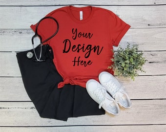 May include: Red t-shirt with the text "Your Design Here" on a white wood background. The shirt is paired with black pants and white sneakers.