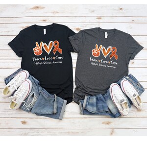 Puede incluir: Dos camisetas con un signo de paz, un coraz&oacute;n y una cinta naranja con el texto "Peace Love Cure Multiple Sclerosis Awareness".