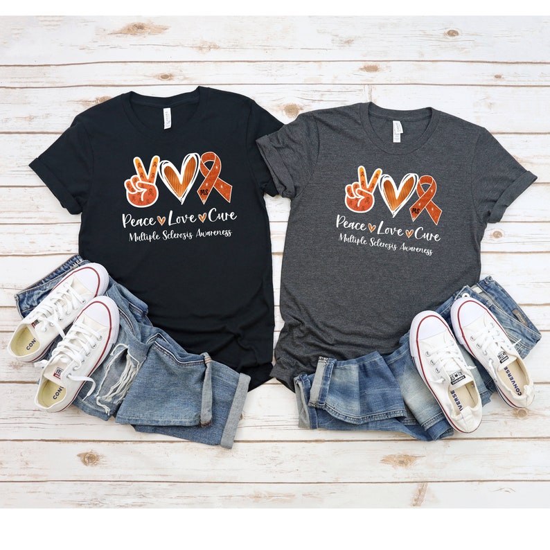Puede incluir: Dos camisetas con un signo de paz, un coraz&oacute;n y una cinta naranja con el texto "Peace Love Cure Multiple Sclerosis Awareness".