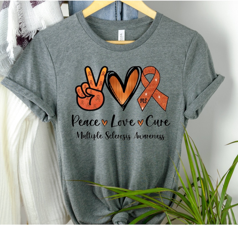 Puede incluir: Camiseta gris con un signo de paz, un coraz&oacute;n y una cinta naranja con el texto "Peace Love Cure" y "Multiple Sclerosis Awareness".