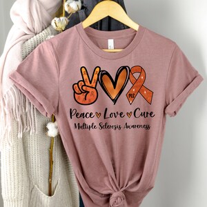 Puede incluir: Una camiseta rosa con un signo de paz, un coraz&oacute;n y una cinta con purpurina naranja y marr&oacute;n. El texto "Peace Love Cure" y "Multiple Sclerosis Awareness" est&aacute; impreso en la camiseta.