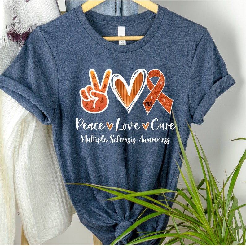 Puede incluir: Una camiseta azul marino con un signo de paz, un coraz&oacute;n y una cinta naranja con el texto "Peace Love Cure" y "Multiple Sclerosis Awareness".